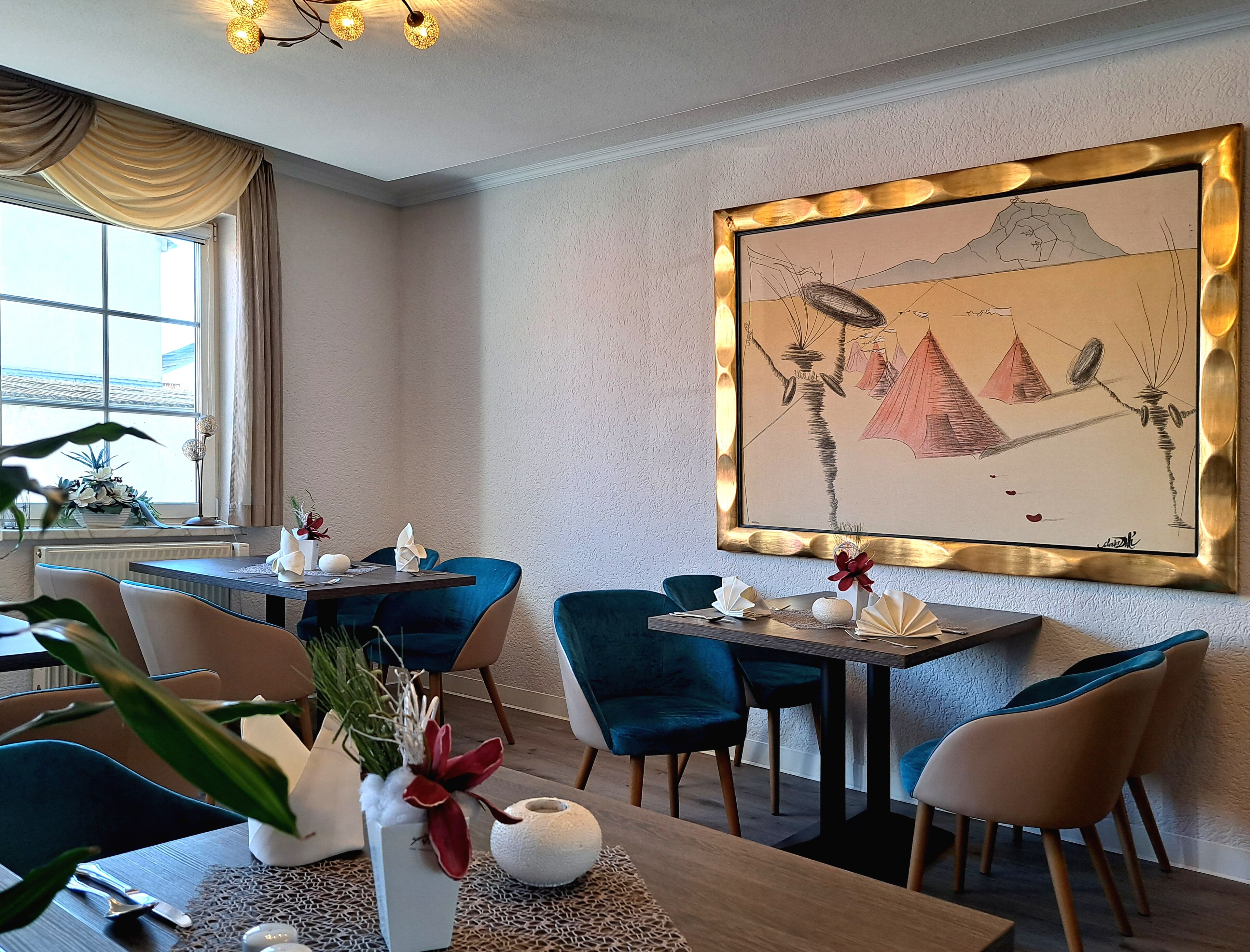A dining area with a table set for two, blue chairs, a vase with red flowers, and a large framed artwork on the wall.