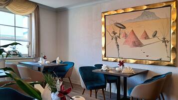 A dining area with a table set for two, blue chairs, a vase with red flowers, and a large framed artwork on the wall.