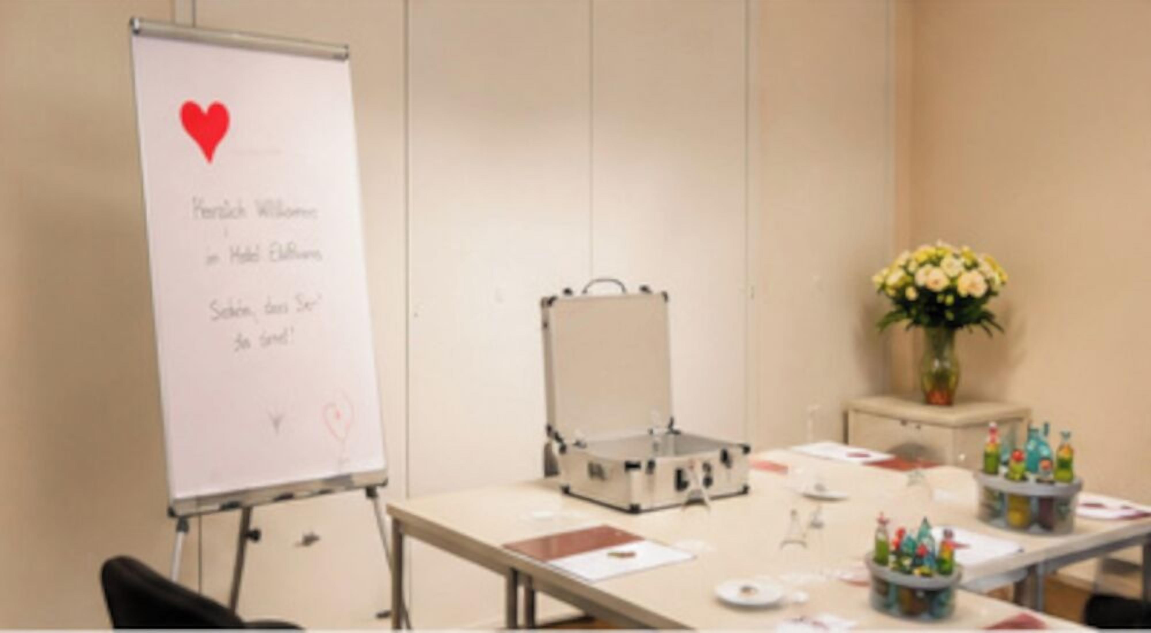 Et mødelokale med en whiteboardtavle, en projektor, et bord med dokumenter og en vase med blomster.