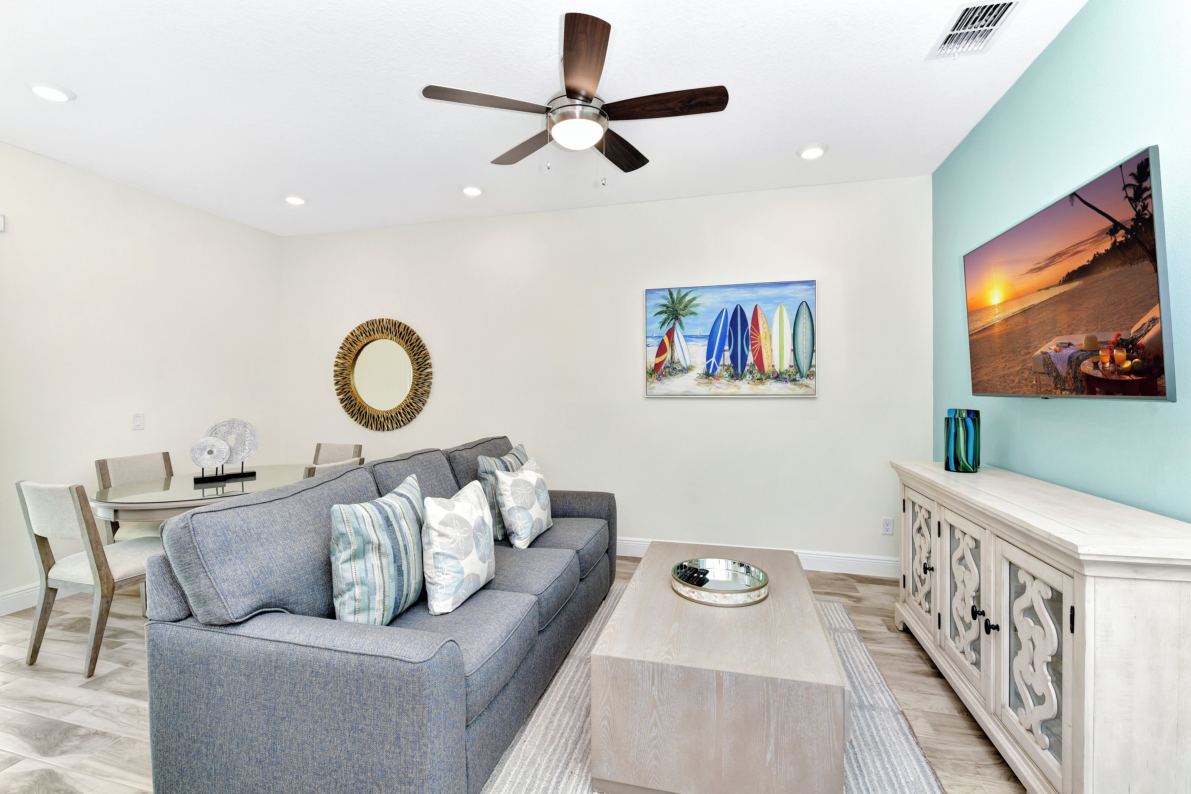 A living room with a gray sofa, a coffee table, a dining table with chairs, a ceiling fan, and wall art.