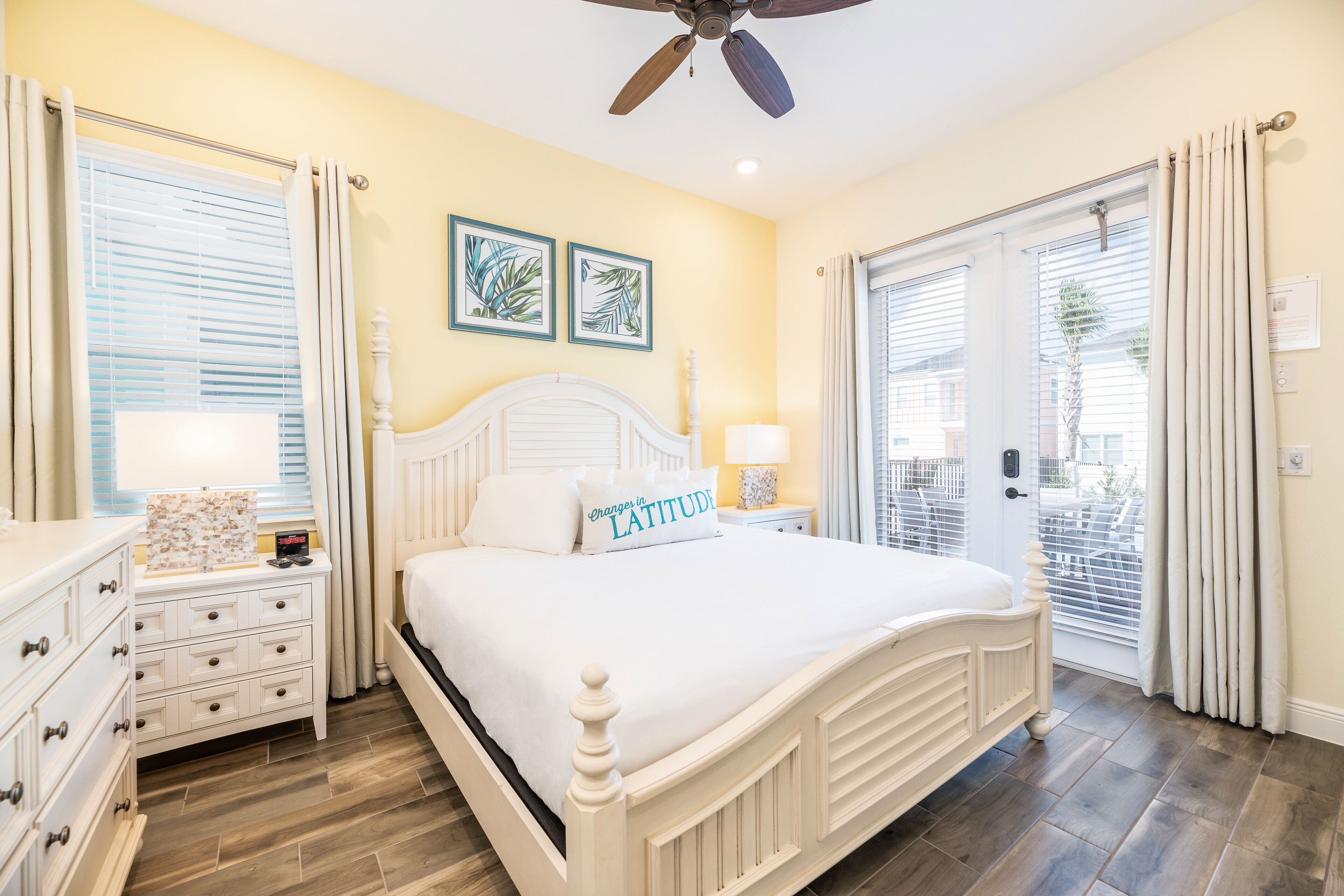 A bedroom with a white bed, two bedside tables with lamps, a ceiling fan, and a view of the outside through a sliding glass door.