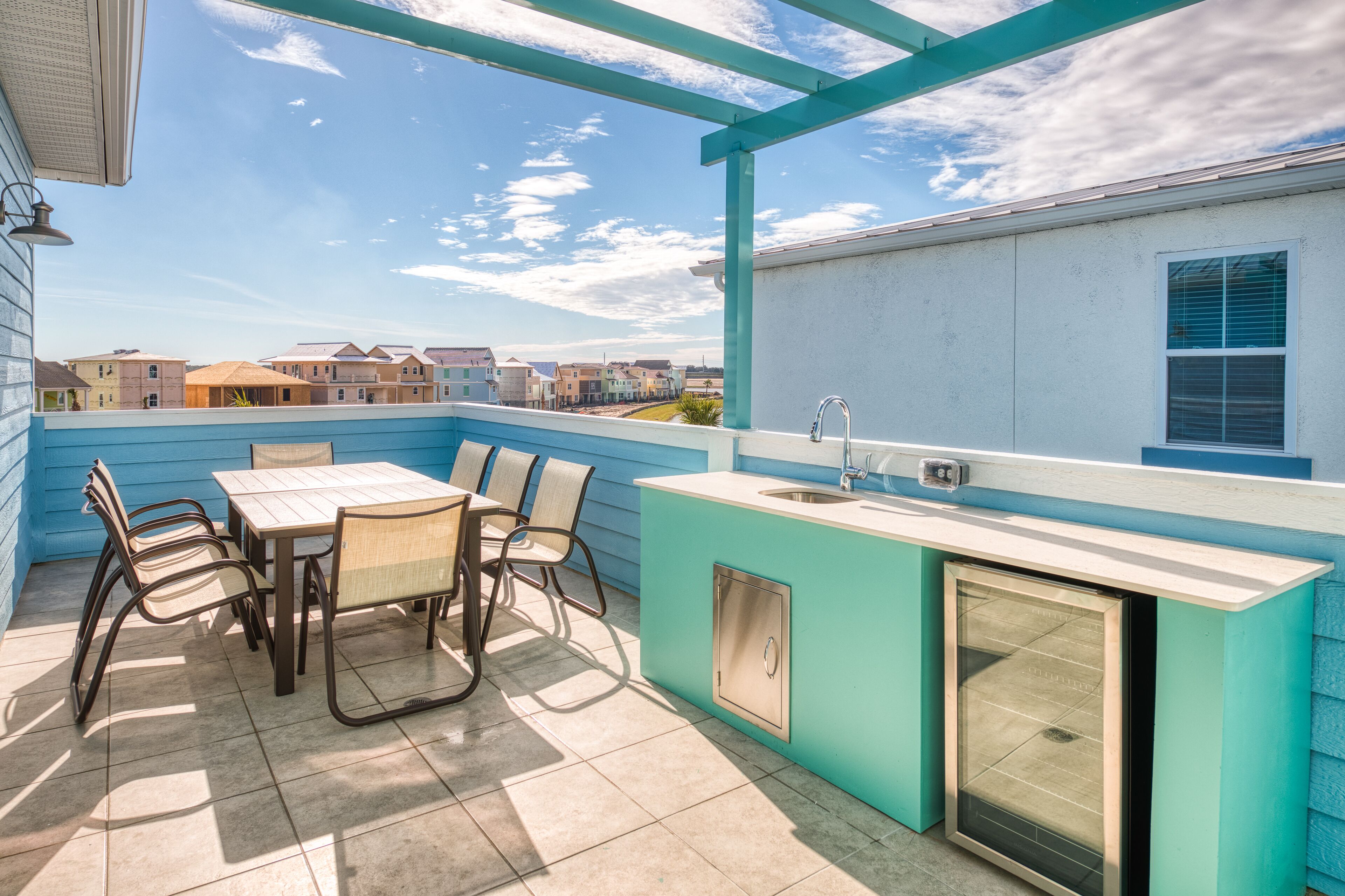 A blue outdoor kitchen with a dining table and chairs, a sink, and a window overlooking a residential area.