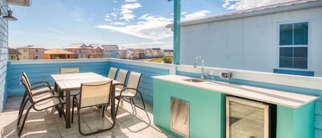 Une cuisine extérieure bleue, avec une table et des chaises pour les repas, un évier et une fenêtre donnant sur un quartier résidentiel.