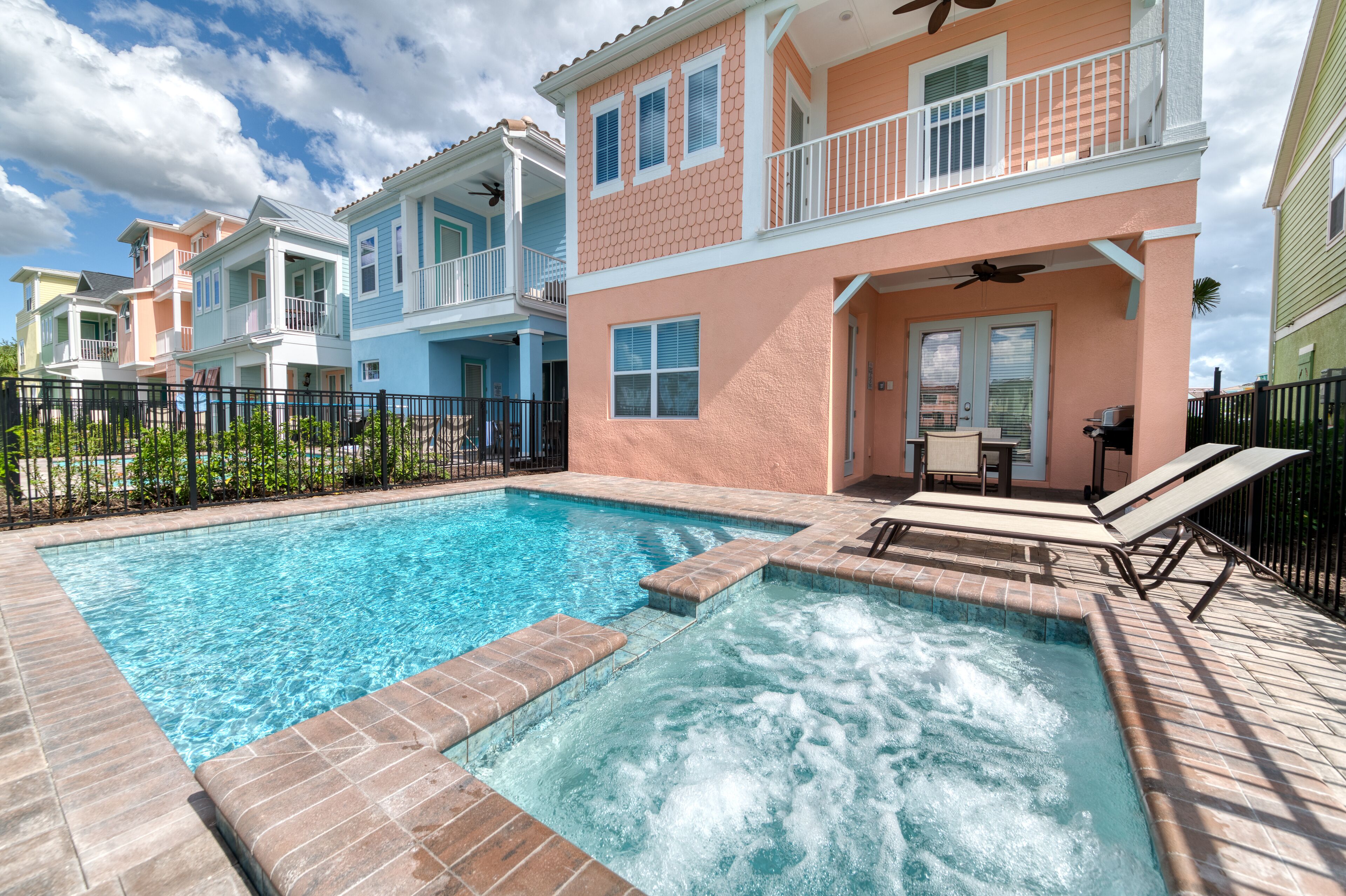 A swimming pool with a hot tub, surrounded by a fence and a row of colorful houses.