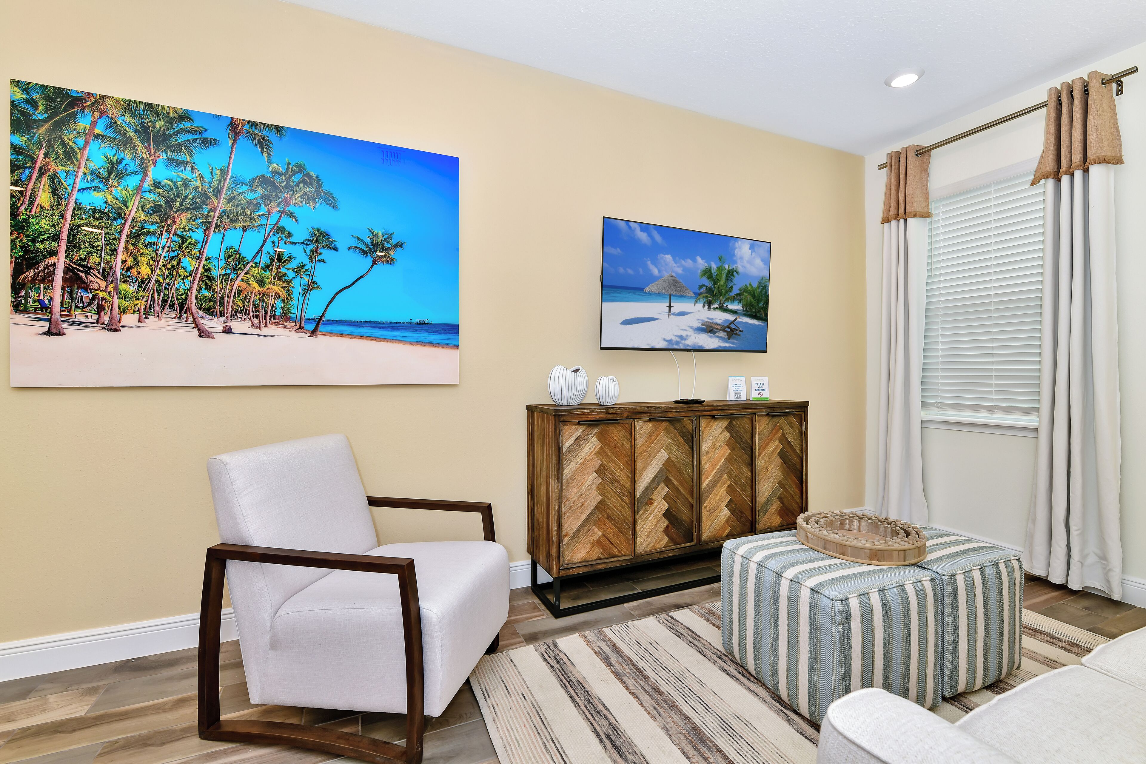 A living room with a white armchair, a striped ottoman, a herringbone-patterned cabinet, and two beach-themed paintings on the wall.