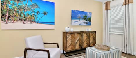 A living room with a white armchair, a striped ottoman, a herringbone-patterned cabinet, and two beach-themed paintings on the wall.