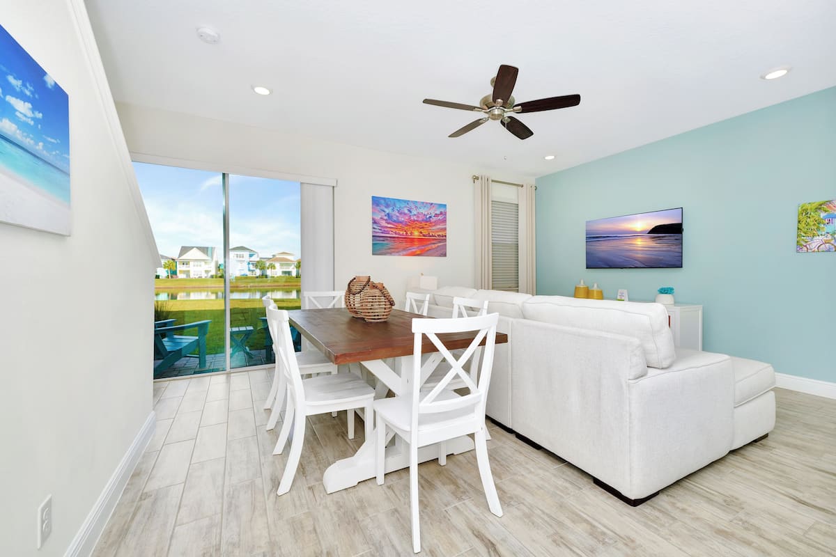 A modern living room with a dining area, a ceiling fan, and large windows overlooking a pool.