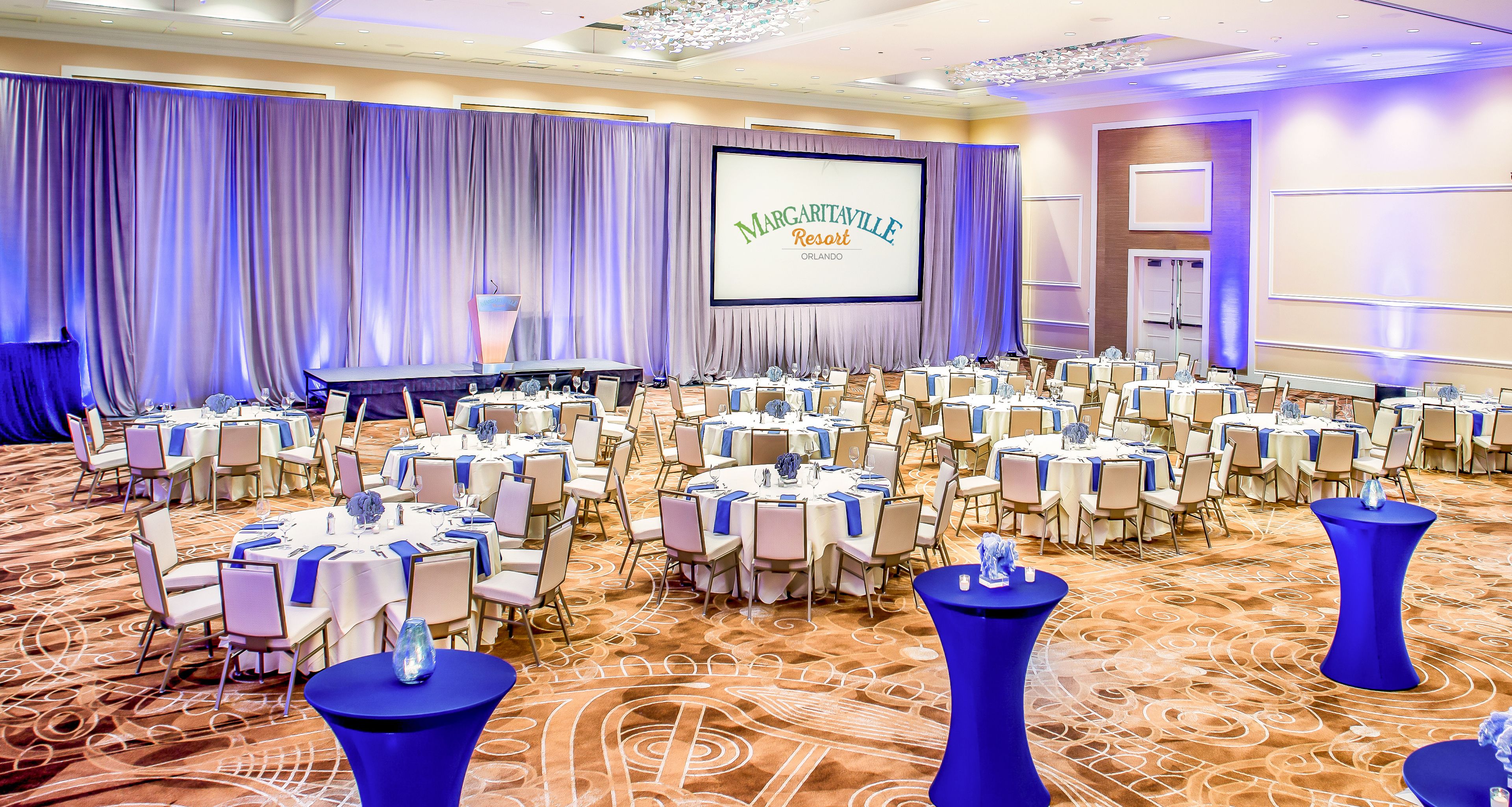 A conference room at MarGaritaville Resort in Orlando with multiple round tables, chairs, and a large screen displaying the resort's name.