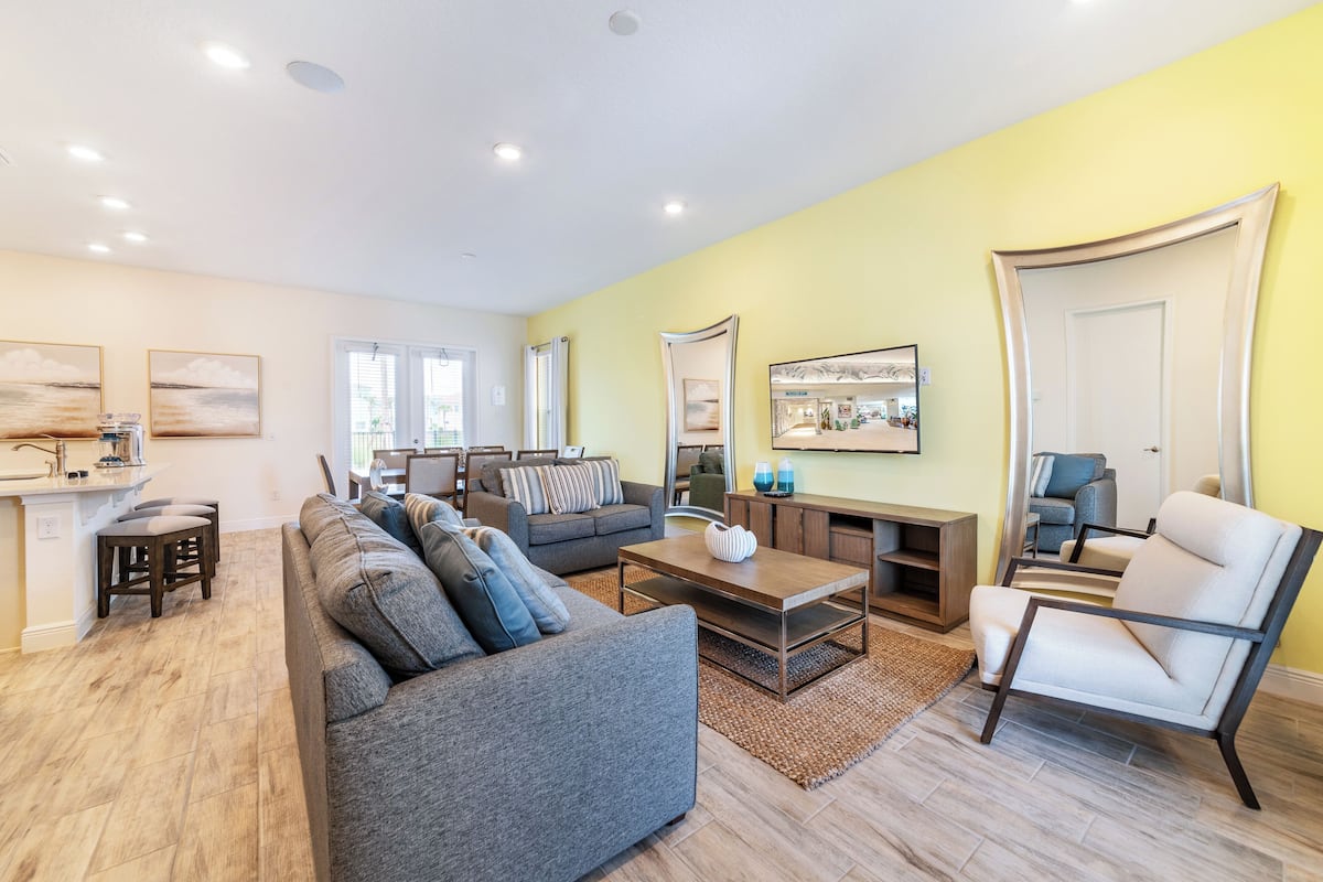 A modern living room with a sofa, armchairs, and a coffee table. There is a dining area with a table and chairs. The room has a yellow wall and a large mirror.