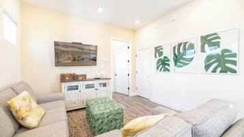 Una sala de estar con un sofá gris, un reposapiés y un mueble bajo. Hay tres cuadros en la pared y una puerta que conduce a otra habitación.