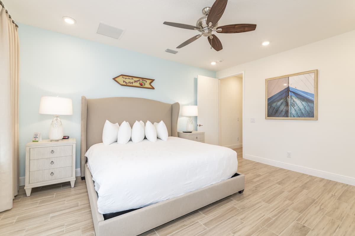 A bedroom with a large bed, two bedside tables with lamps, a ceiling fan, a wall-mounted artwork, and a door leading to another room.