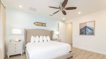 Un dormitorio con una cama grande, dos mesitas de noche con lámparas, un ventilador de techo, una obra de arte en la pared y una puerta que da a otra habitación.