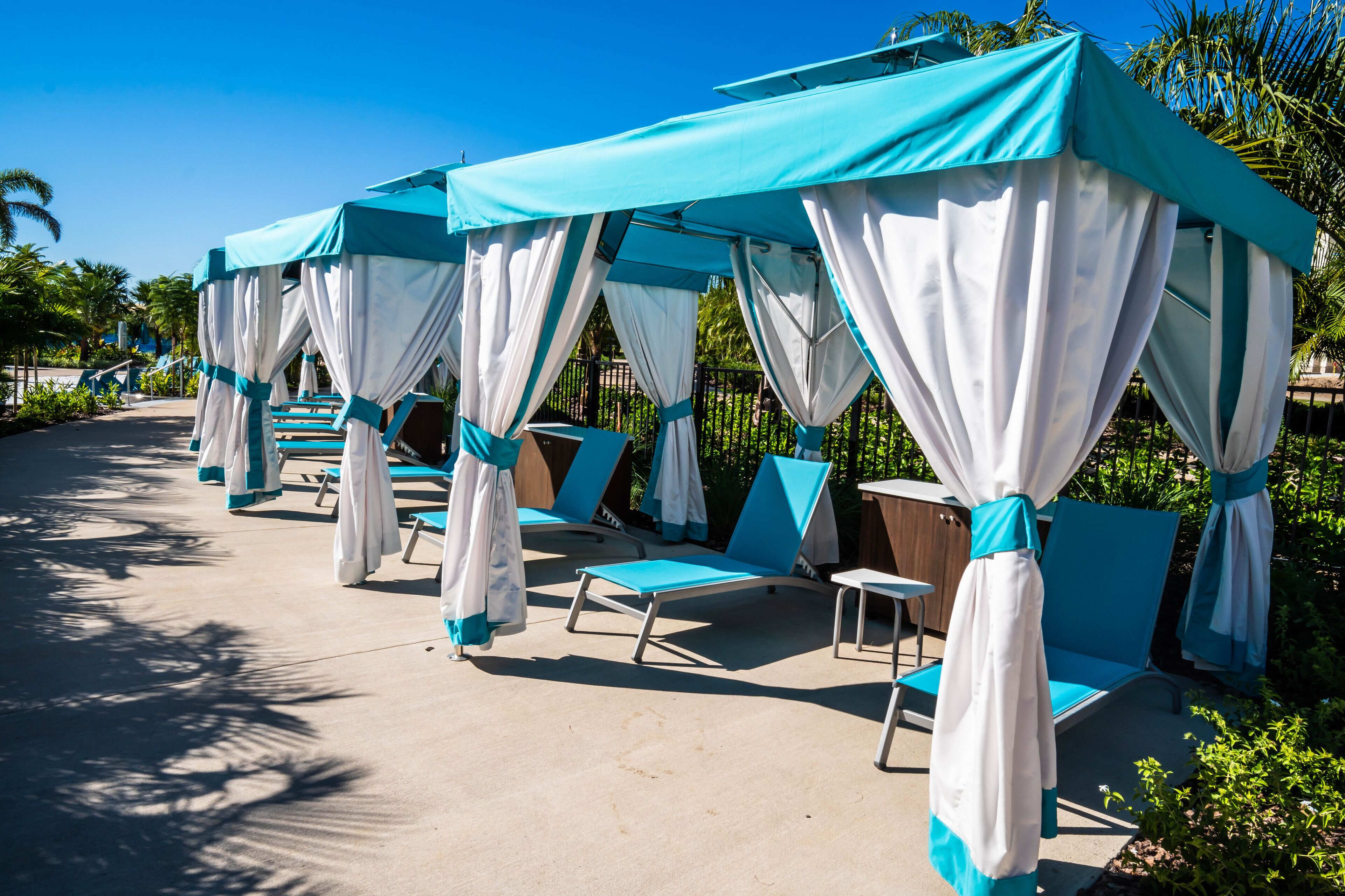 A series of covered lounge areas with blue and white canopies, arranged along a pathway with palm trees and greenery in the background.