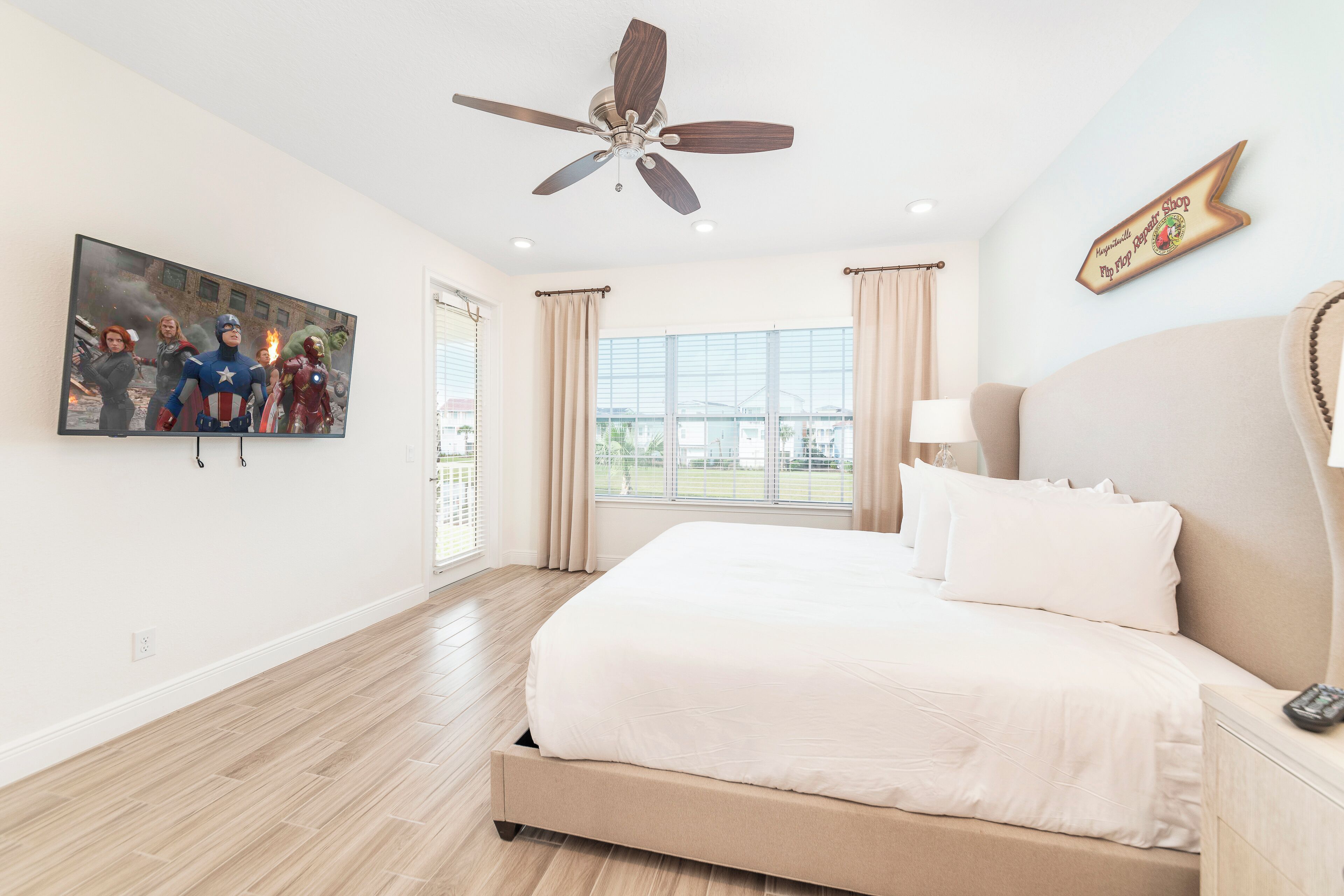 A bedroom with a large bed, a ceiling fan, a TV on the wall, and a window with curtains.