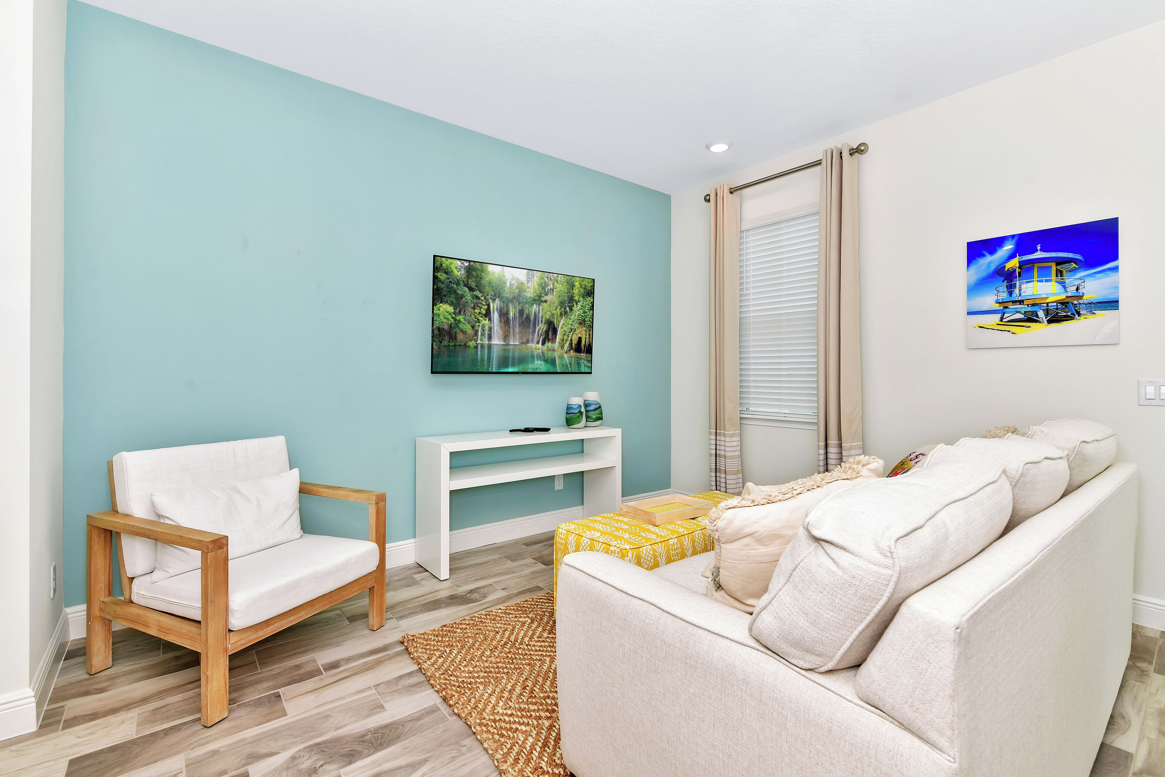 A living room with a teal wall, a white sofa, a wooden chair, a white console table, and a TV on the wall.