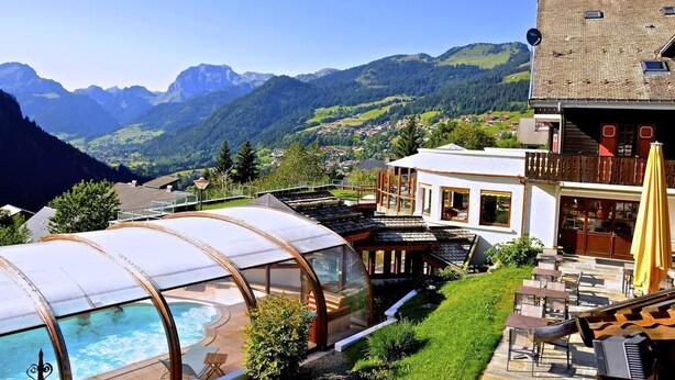 A modern hotel with a glass-enclosed pool, a wooden deck, and a view of mountains. - Fleur De Neige Hôtel (Chatel)