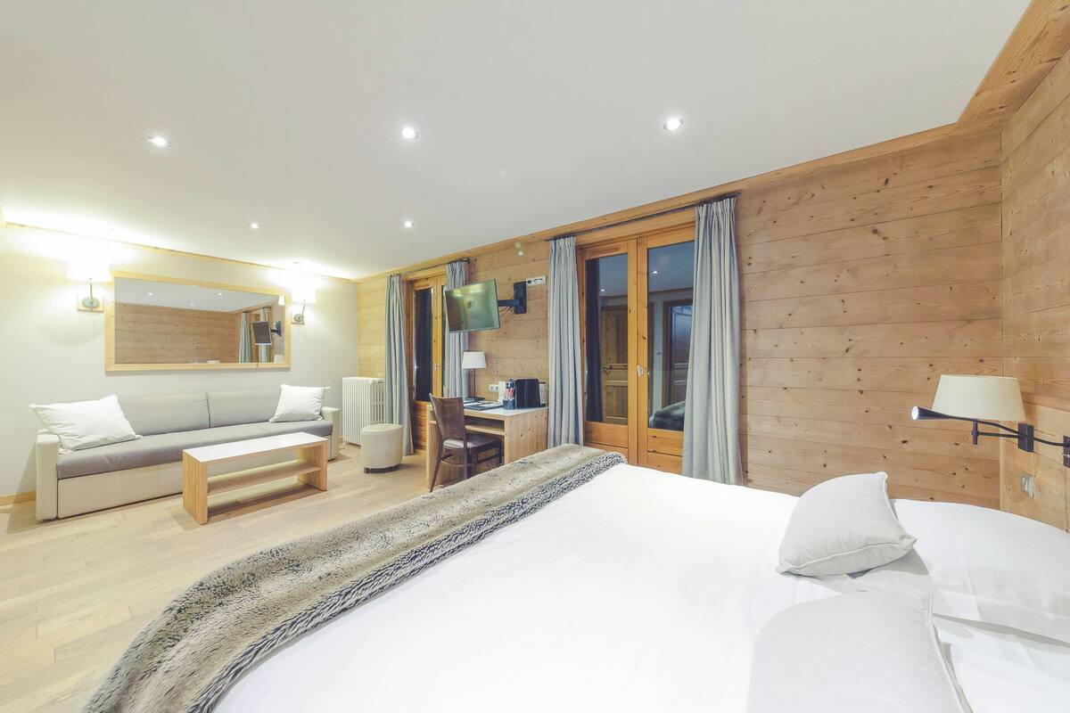 A modern hotel room with a wooden accent wall, a large bed, a sofa, a desk, and a TV.