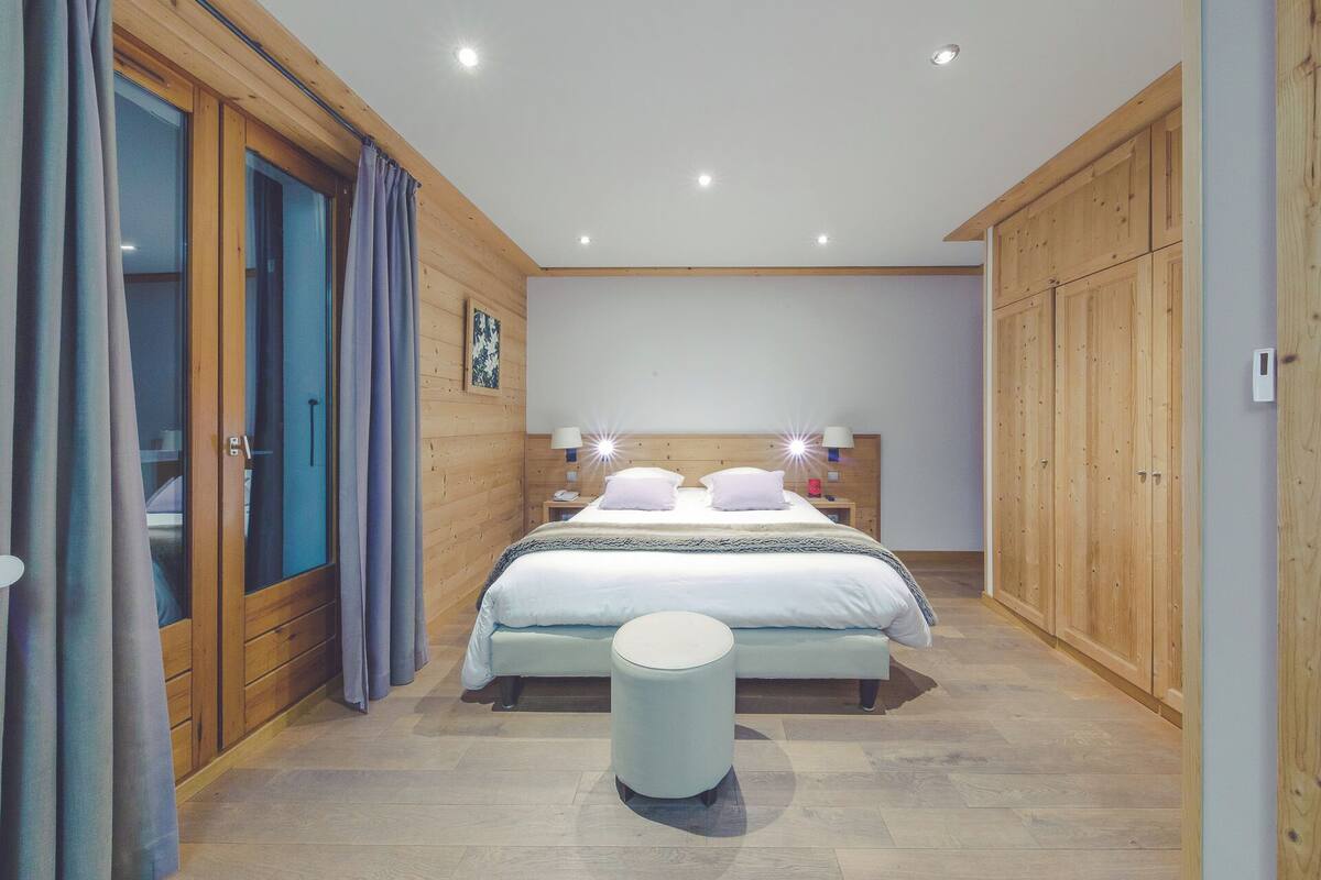 A bedroom with a bed, bedside tables, a nightstand, a round stool, wooden paneling, and a large wardrobe.