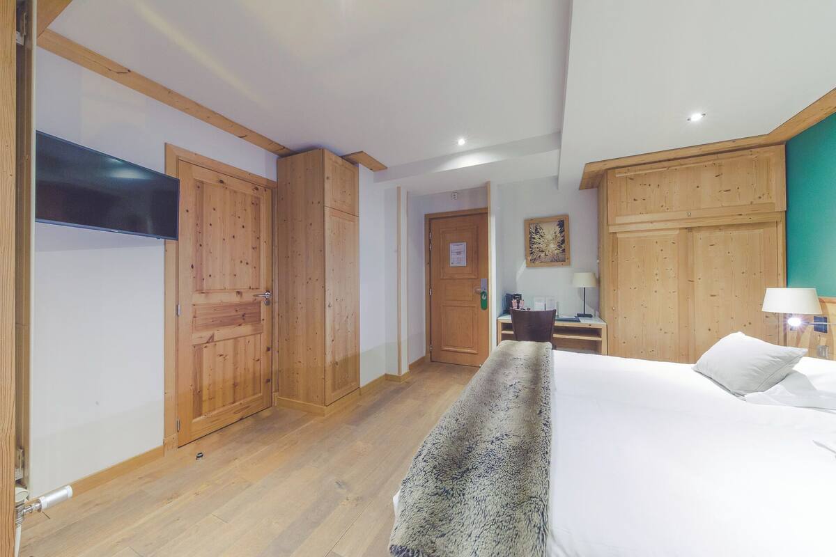 A hotel room with wooden wardrobes, a bed with white linens, a desk with a lamp, and a television on the wall.