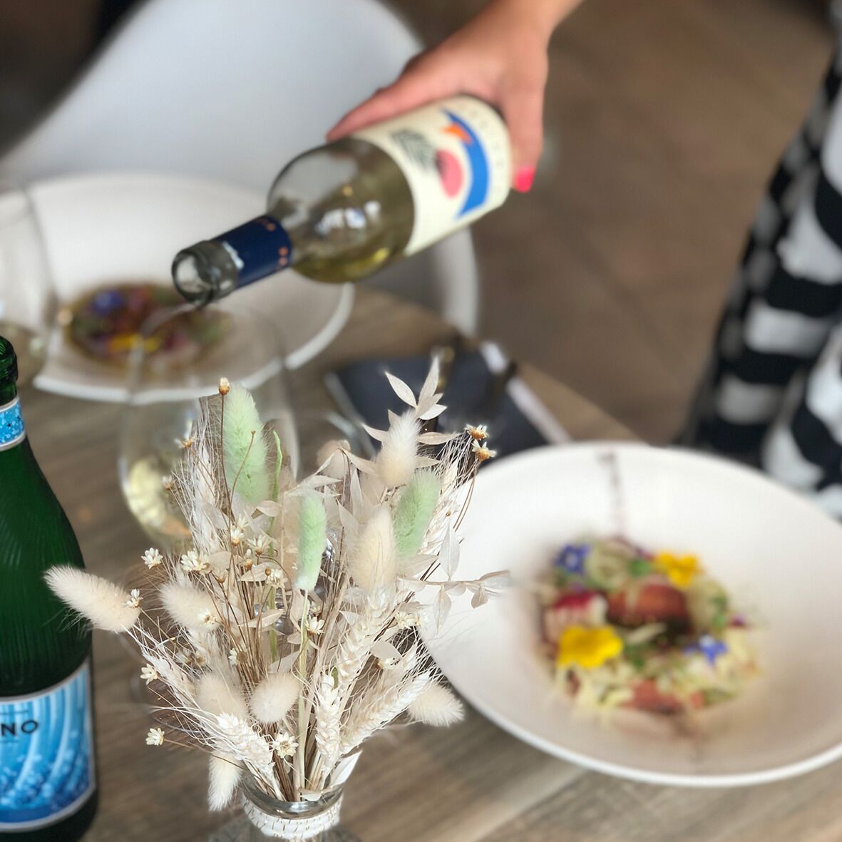 Uma garrafa de vinho sendo servida em uma taça, uma saladeira e um vaso com flores secas sobre uma mesa.