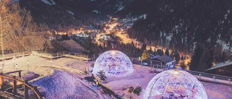 A snow-covered landscape with two illuminated geodesic domes, a wooden deck, and a view of a village in the background.