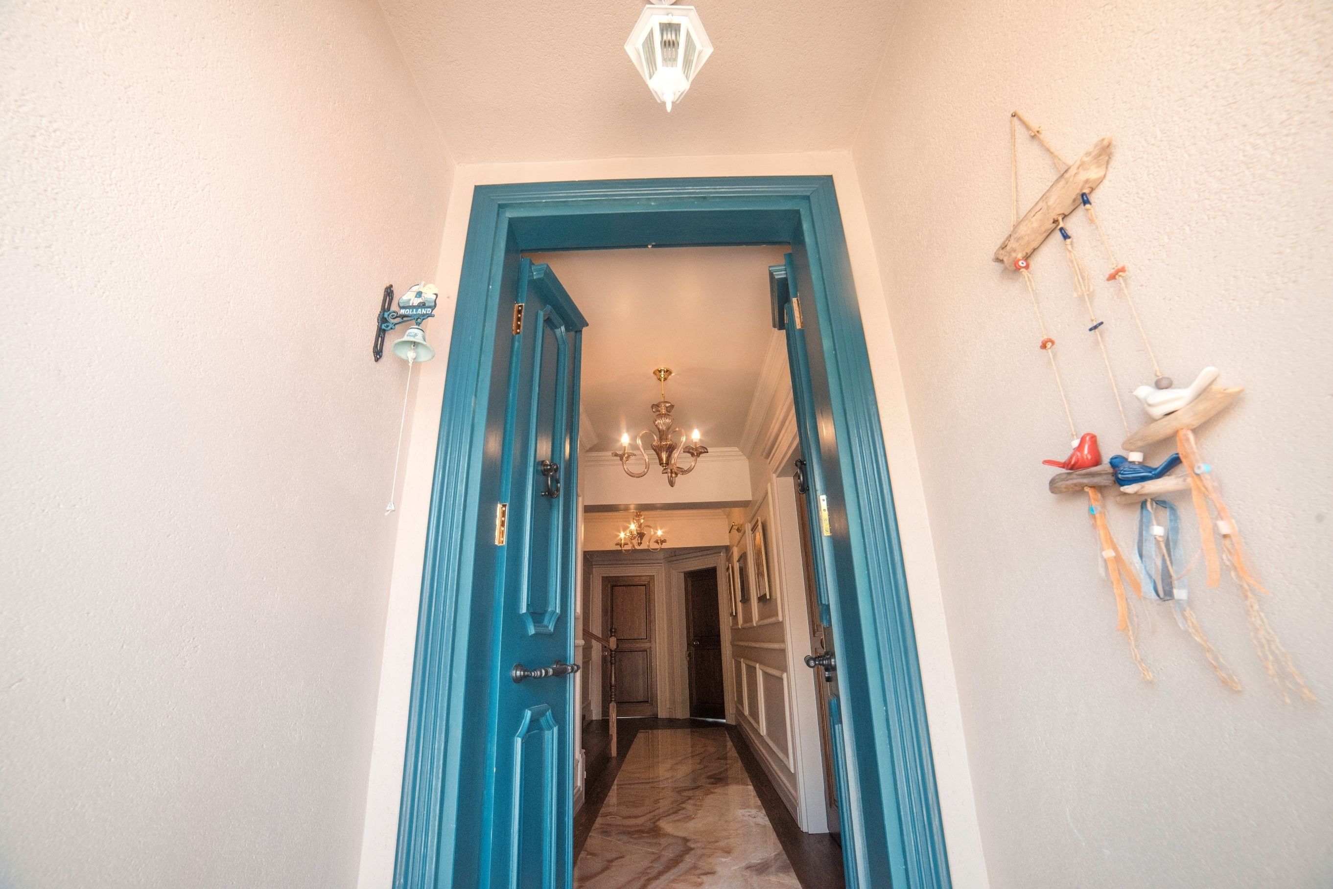 Corredor com porta azul que dá para um quarto. É possível ver um candelabro e elementos decorativos nas paredes.