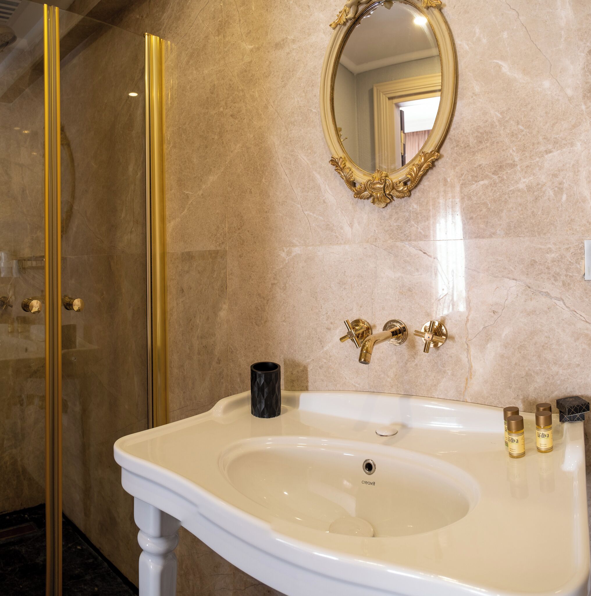 Un baño con pared de mármol, espejo con marco dorado, lavamanos blanco e grifos dorados.