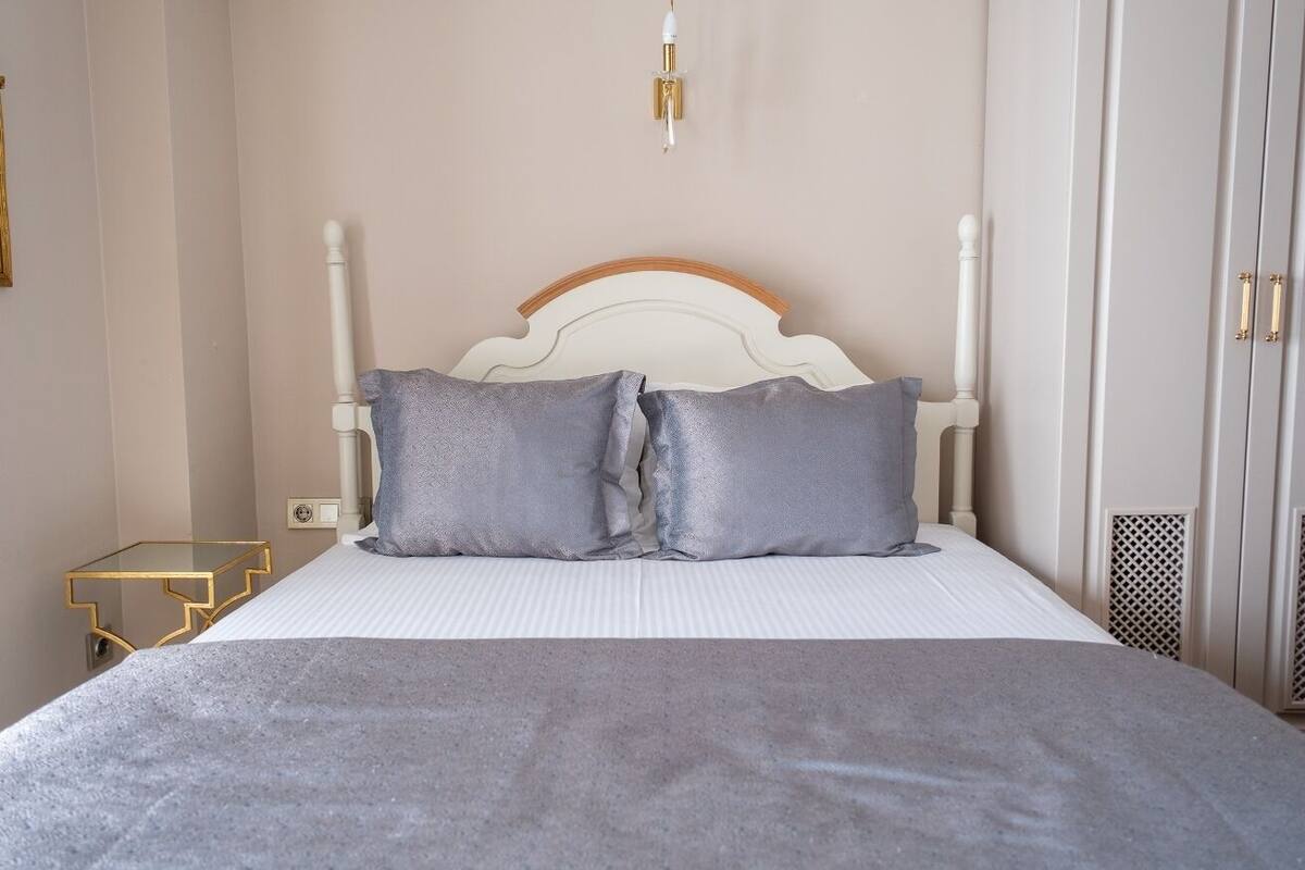 A neatly made bed with a white headboard and two grey pillows, a small bedside table with a gold frame, and a wall-mounted light fixture.