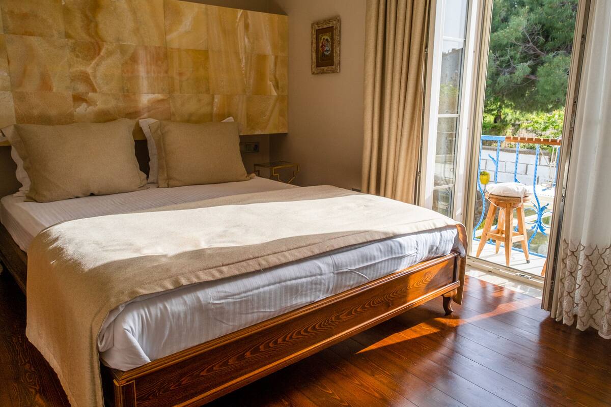 A bedroom with a wooden bed, a headboard, a nightstand, a window with curtains, and a view of greenery outside.