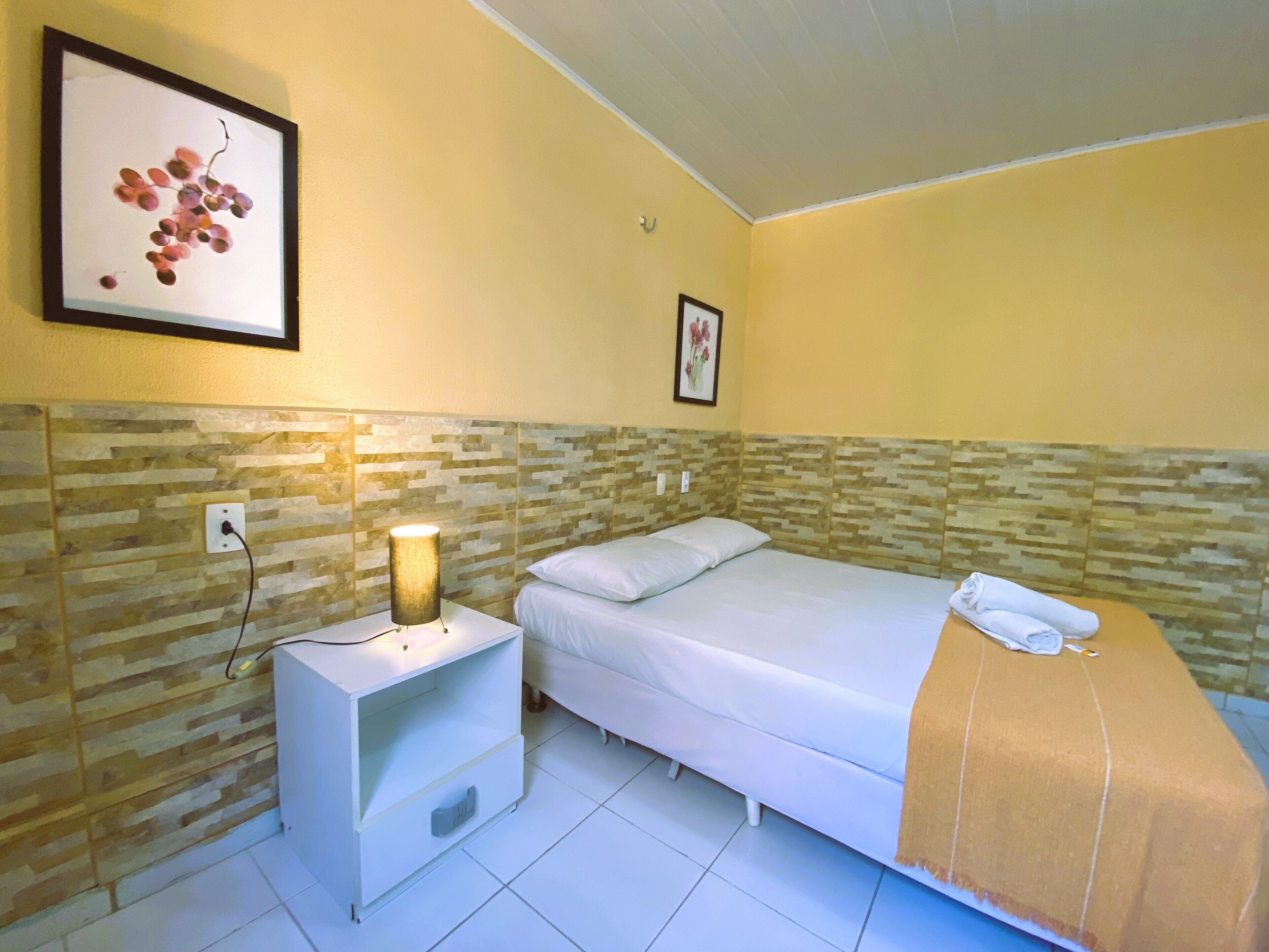 Quarto de hotel com uma cama, mesa de cabeceira com abajur, quadro na parede e chão ladrilhado.