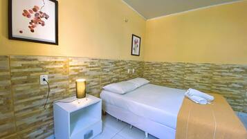 Quarto de hotel com uma cama, mesa de cabeceira com abajur, quadro na parede e chão ladrilhado.