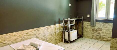 A hotel room with a bed, a small table with towels, a shelf unit, and an air conditioning unit.