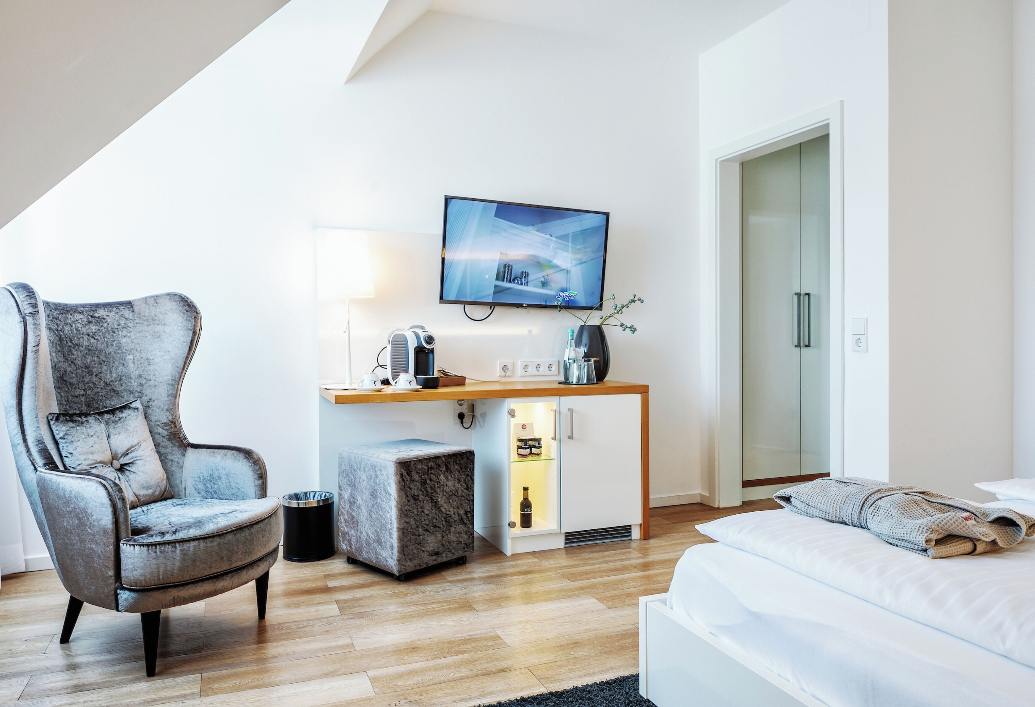 A modern hotel room with a grey armchair, a wooden desk with a flat-screen TV, a minibar, and a bed with a white comforter.