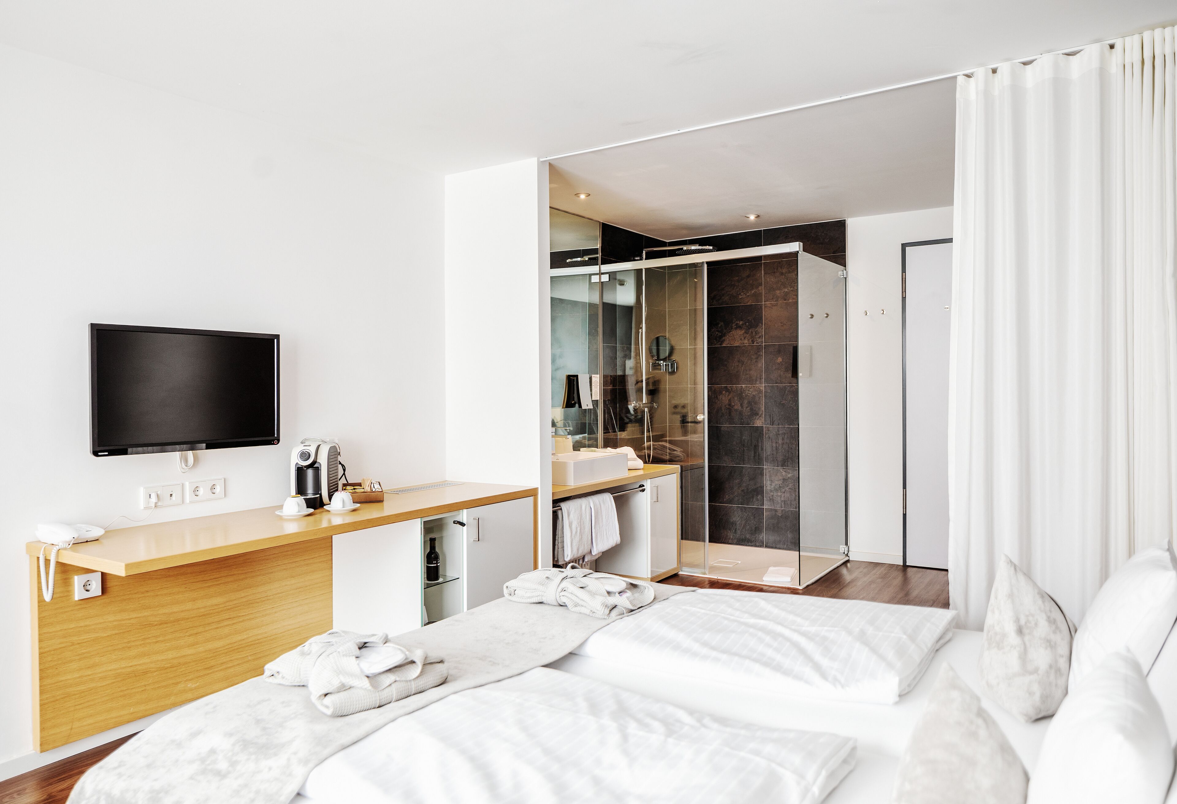 A modern hotel room with a bed, a desk with a flat-screen TV, a shower area, and a bathroom.