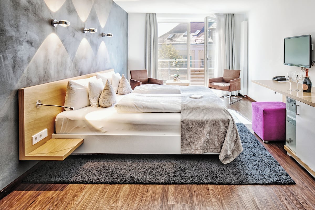 A modern hotel room with a large bed, a desk, a TV, and a balcony with a view.