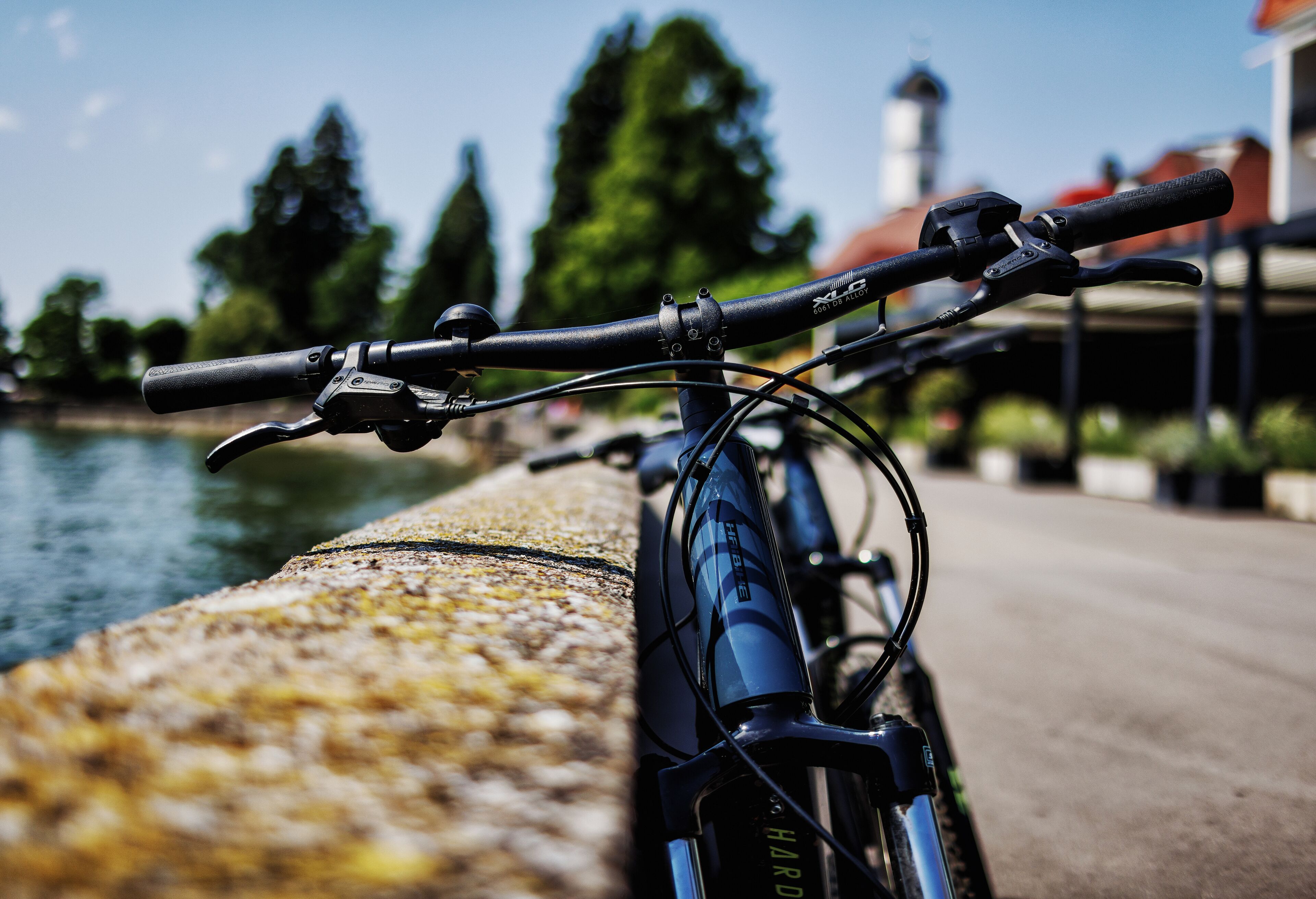 A bicycle parked by a waterfront with a stone railing, a clear sky, and buildings in the background.