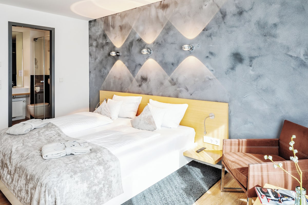 A modern hotel room with a large bed, a wooden nightstand, a seating area with a brown leather sofa, and a wall-mounted light fixture.