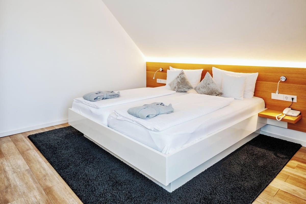 A modern hotel room with a neatly made bed, a wooden headboard, a bedside table with a phone, and a dark rug.