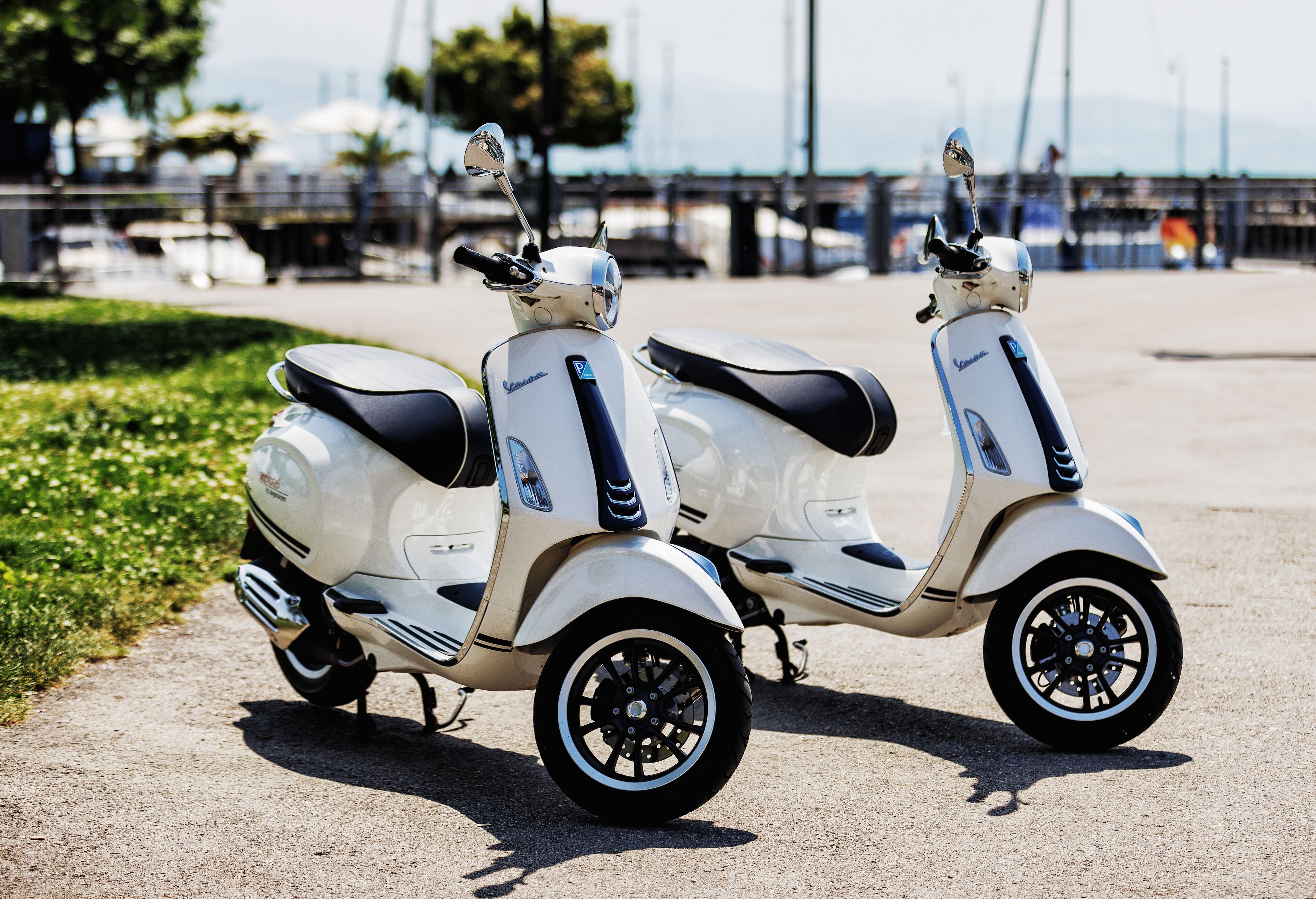 Two white scooters parked on a paved area with a grassy patch and a railing in the background.