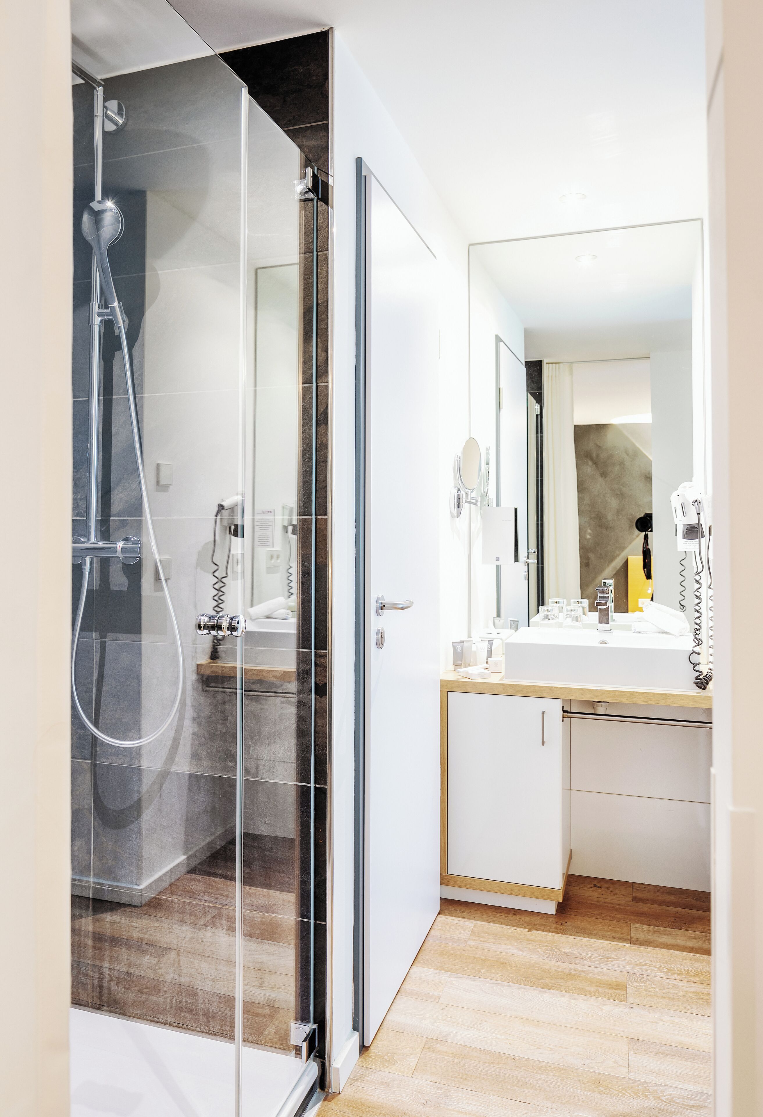 A modern bathroom with a glass shower enclosure, a white sink with a mirror, and a wooden floor.