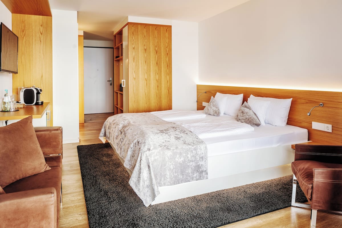 A modern hotel room with a large bed, a wooden wardrobe, a seating area with a brown sofa, and a small table with a kettle.