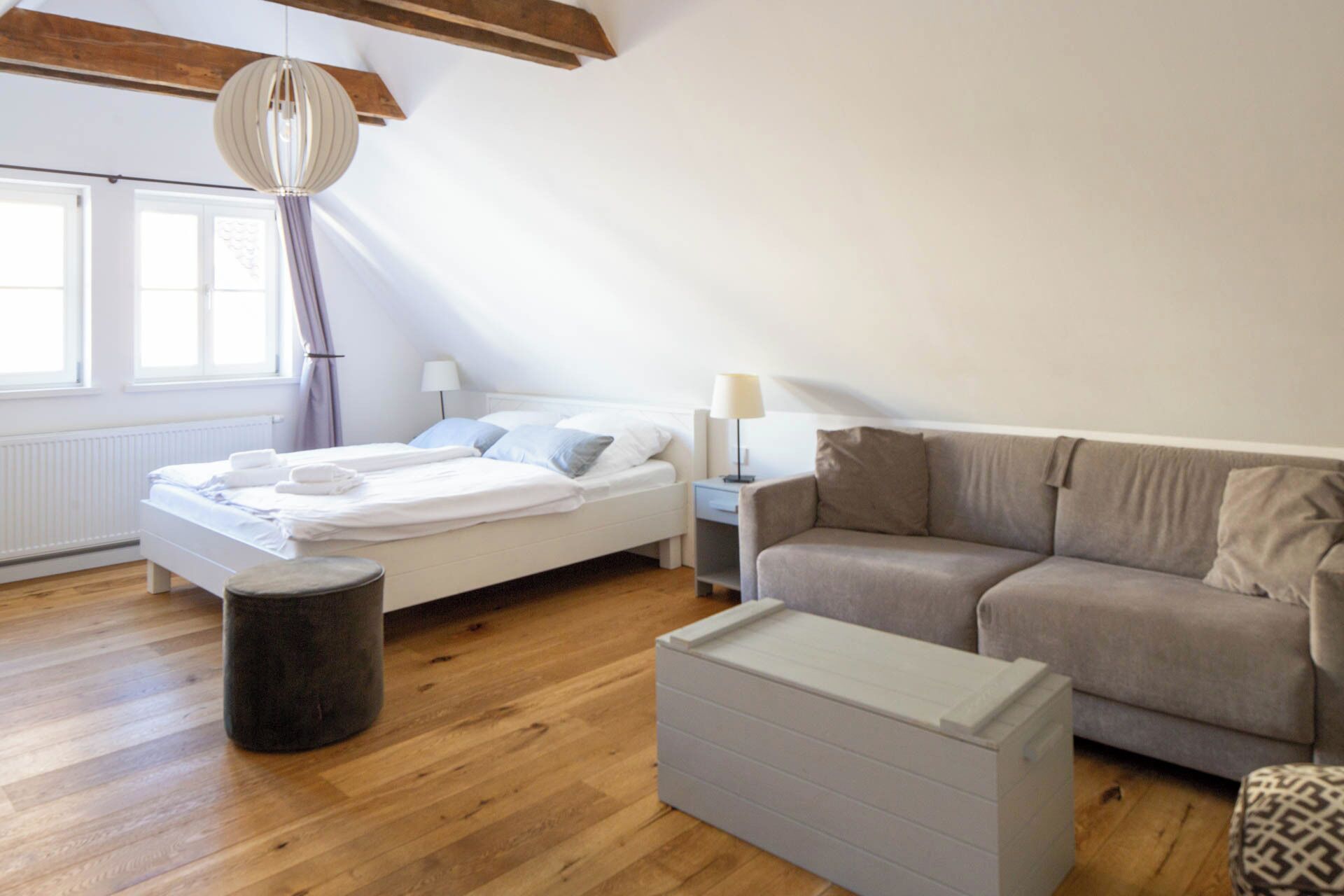 A modern, well-lit room with a wooden floor, a bed with white bedding, a grey sofa, and a small white cabinet.
