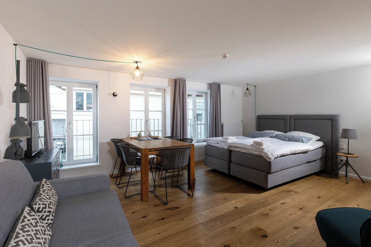 A modern, well-lit room with a wooden floor, a grey sofa, a bed with grey bedding, a dining table with chairs, and a hanging light fixture.