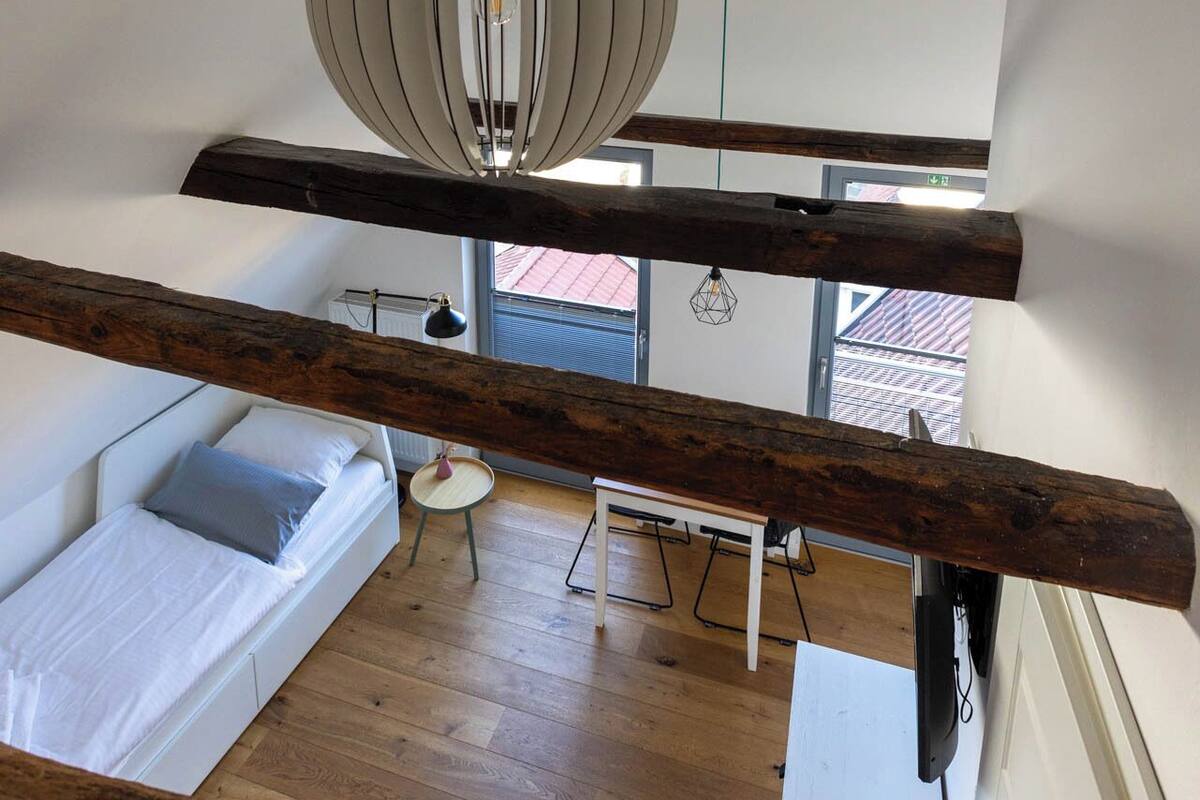 A loft-style bedroom with a sloped ceiling, a hanging light fixture, a bed, a small table, and a view of the outside.