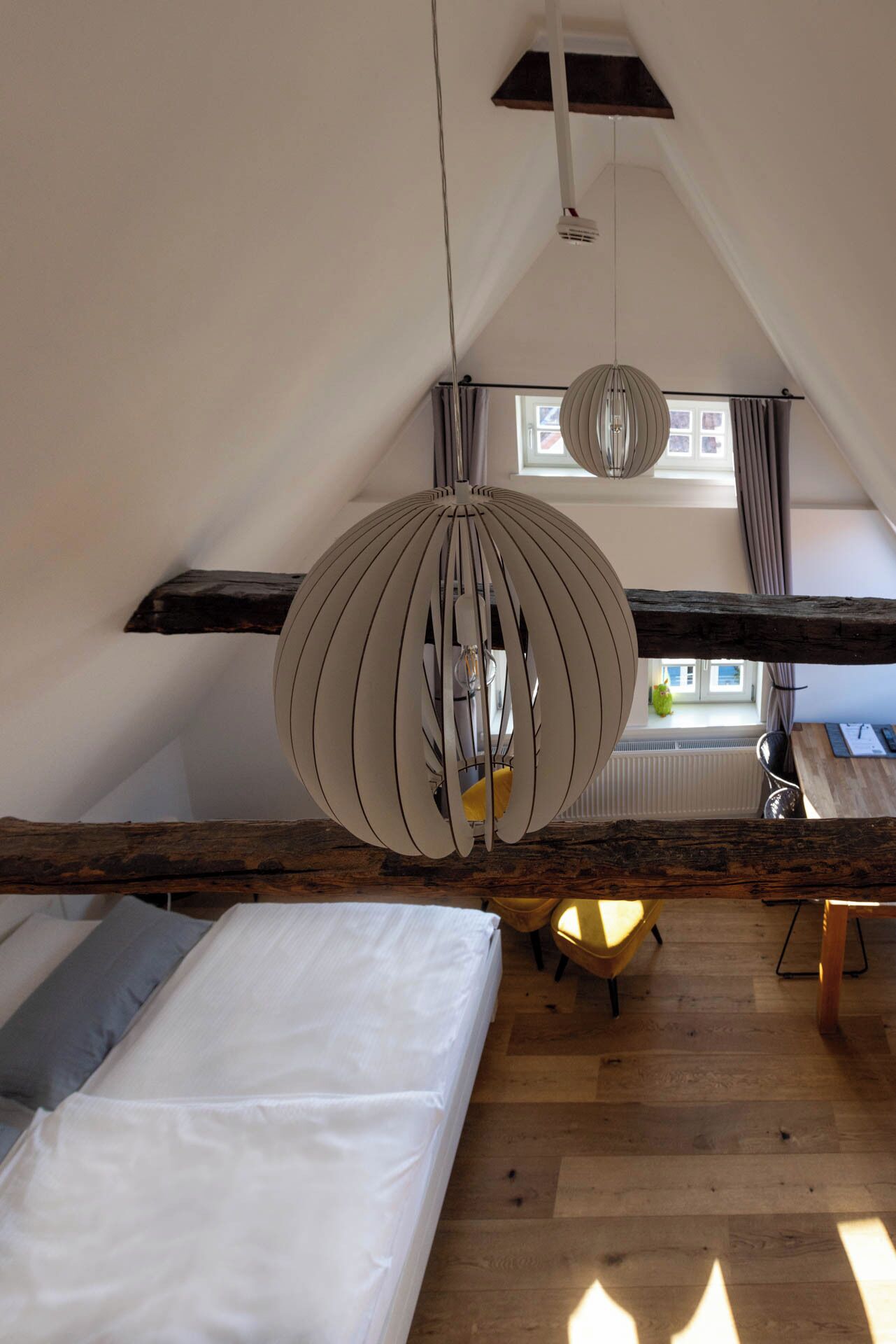 A loft bedroom with a sloped ceiling, a wooden beam ceiling, a bed, a desk, and a hanging light fixture.