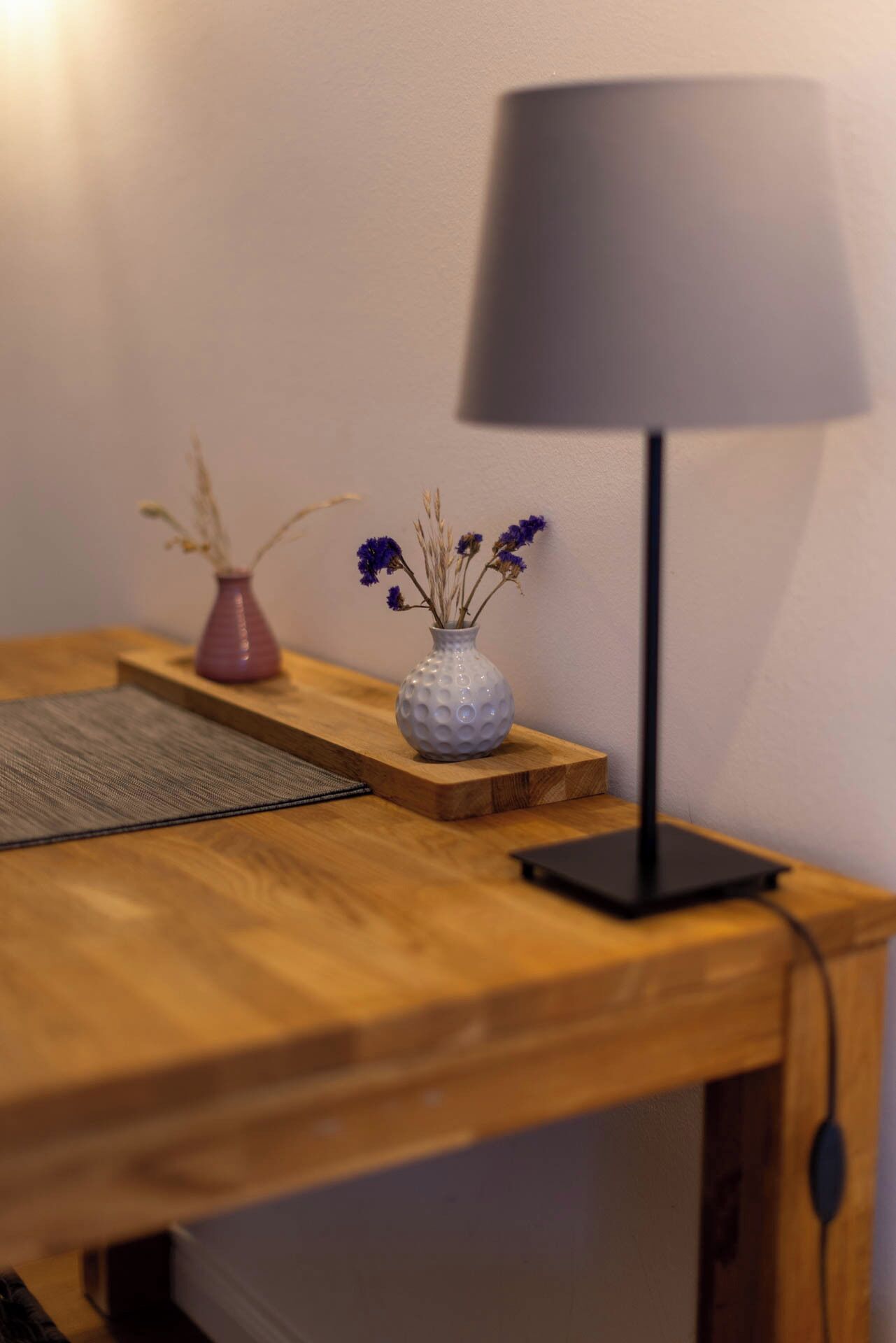 A wooden desk with a white vase containing dried flowers, a small pink vase, and a black lamp.