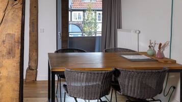 Un comedor con mesa de madera y sillas de mimbre, un radiador y vistas a un edificio a través de una ventana.