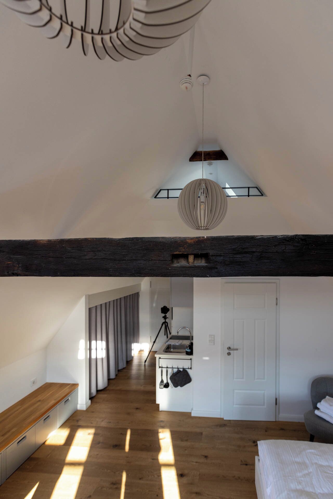 Et rummeligt loftværelse med skrå vægge, synlige træbjælker, et ovenlys og en moderne lysekrone.