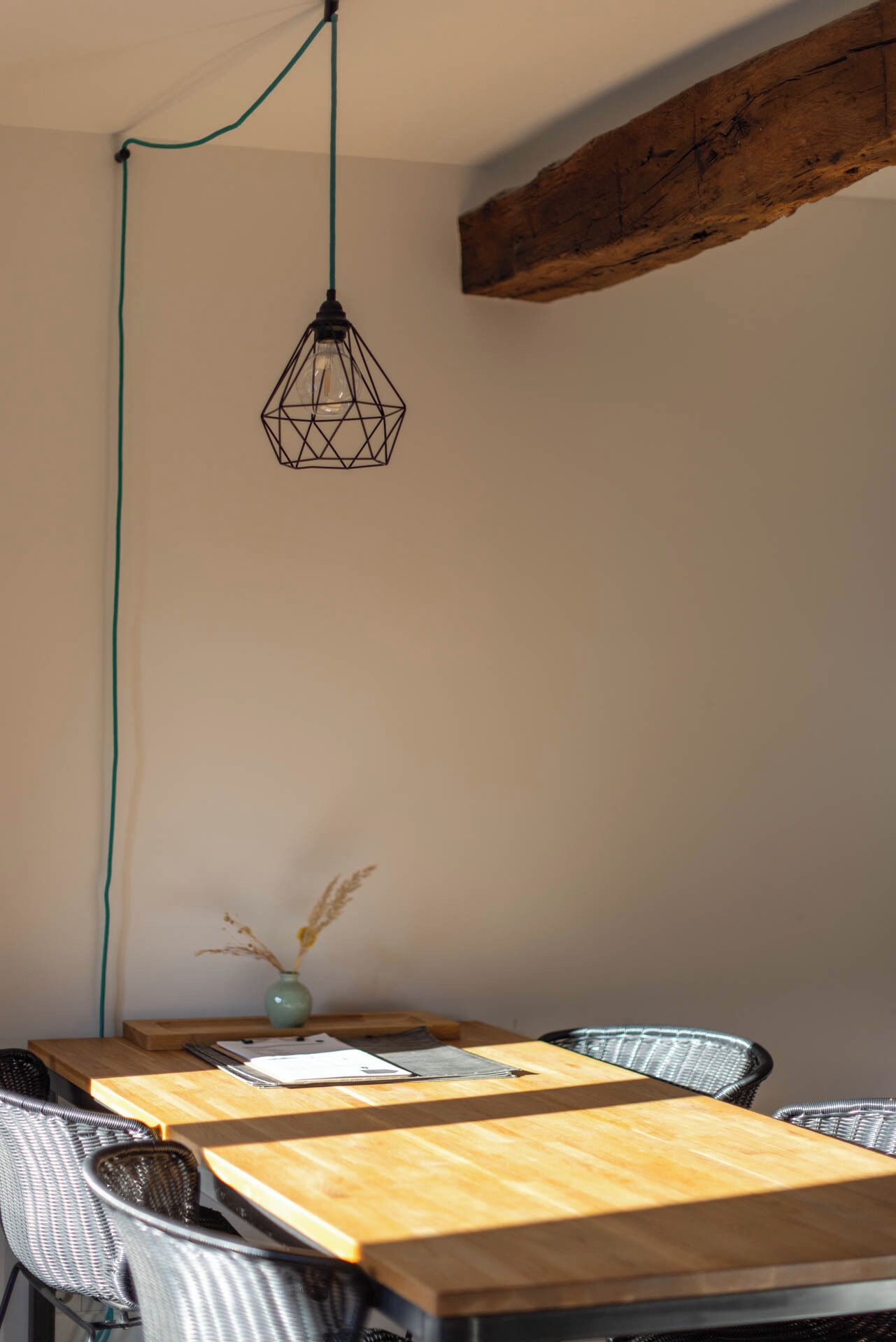 A dining area with a wooden table, wicker chairs, a vase with dried plants, and a geometric pendant light.
