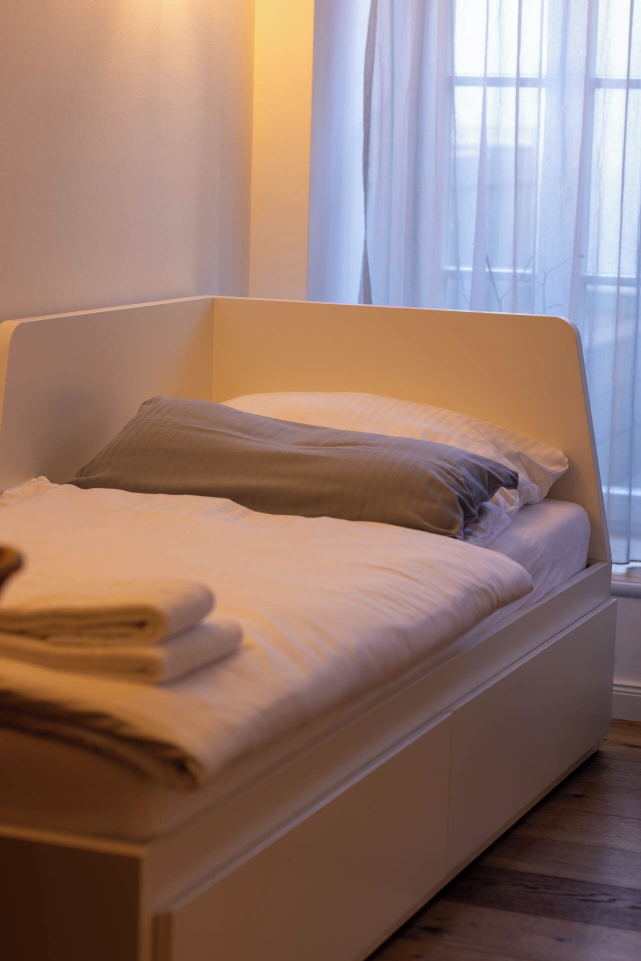 Un lit bien fait, avec du linge de lit blanc et une serviette pliée, une tête de lit blanche et une fenêtre ornée de rideaux légers.